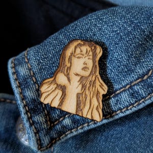 May include: A light-colored wooden pin featuring a laser-engraved portrait of a person with long, wavy hair and defined facial features. The pin is shaped to follow the outline of the figure and is attached to a blue denim fabric surface.