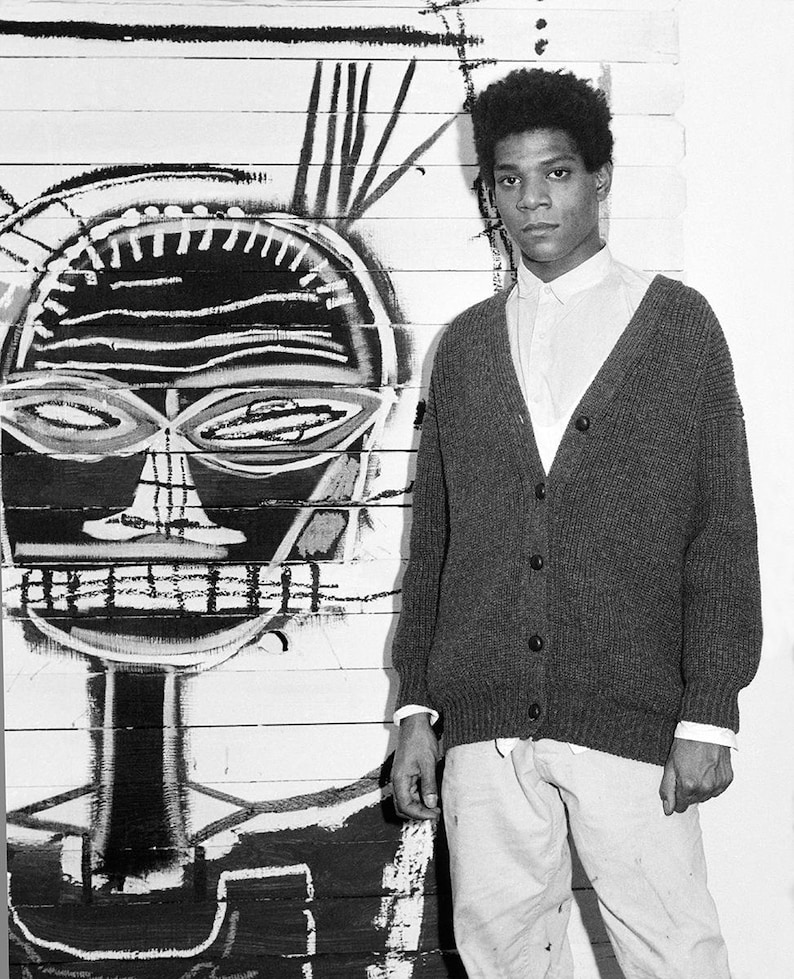 May include: A black and white photo of a man standing in front of a large painting. The painting is a black and white abstract portrait of a person. The man is wearing a dark cardigan sweater and light-colored pants.