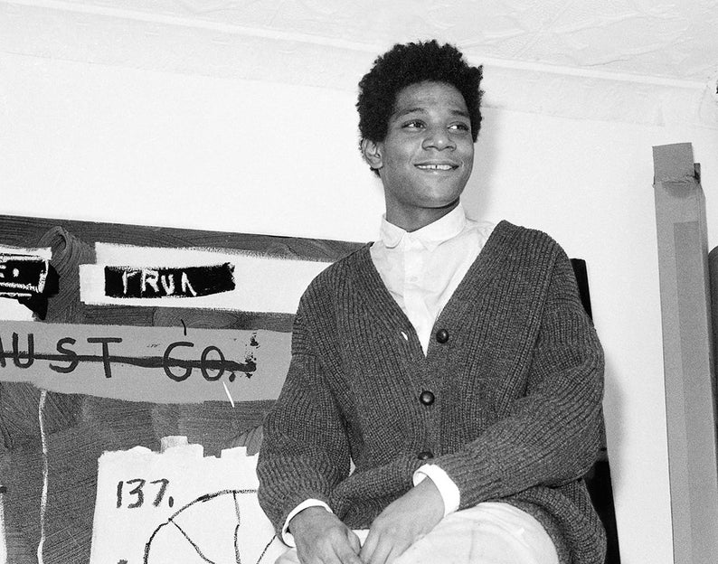 May include: A black and white photo of a man wearing a gray cardigan sweater and a white collared shirt. He is standing in front of a wall with a painting on it. The painting has the words "TRUST" and "MUST GO" written on it.