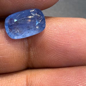 Puede incluir: Una gema azul claro facetada con forma rectangular. La gema, sostenida entre los dedos, presenta fracturas internas. El color de la piedra es un azul claro y vibrante, con sutiles variaciones tonales.