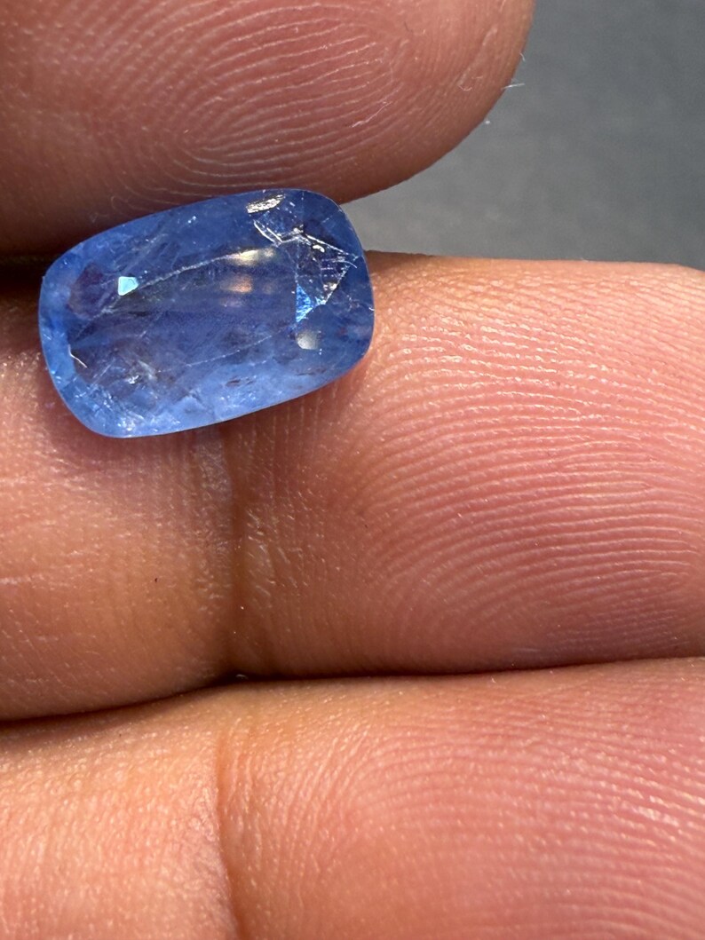 Puede incluir: Una gema azul claro facetada con forma rectangular. La gema, sostenida entre los dedos, presenta fracturas internas. El color de la piedra es un azul claro y vibrante, con sutiles variaciones tonales.