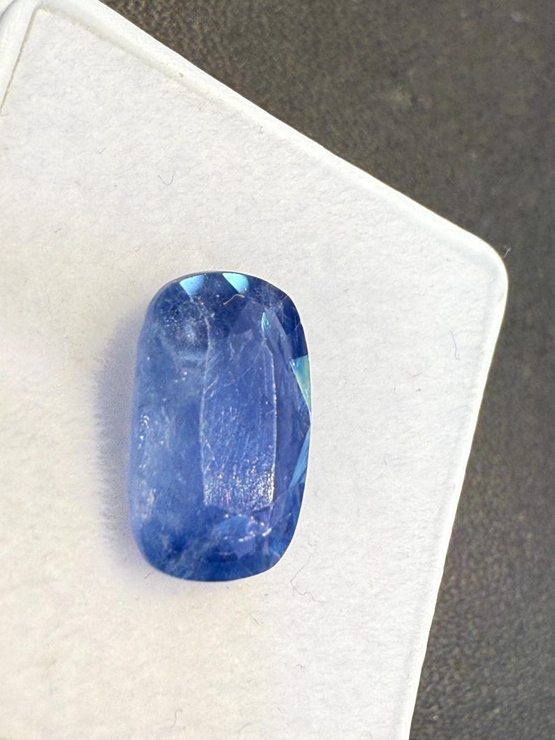 Puede incluir: Una gema azul facetada, de forma ovalada. La gema es de un azul profundo y saturado con un ligero brillo, sobre un fondo blanco. El corte de la piedra crea m&uacute;ltiples facetas que reflejan la luz.