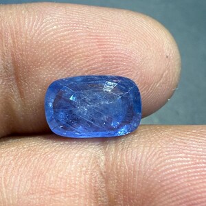 Puede incluir: Una gema azul facetada, de talla coj&iacute;n. La gema se sostiene entre los dedos, mostrando su calidad brillante y transparente. Las facetas de la piedra capturan la luz, creando un efecto brillante. El color general es un azul profundo y vibrante.