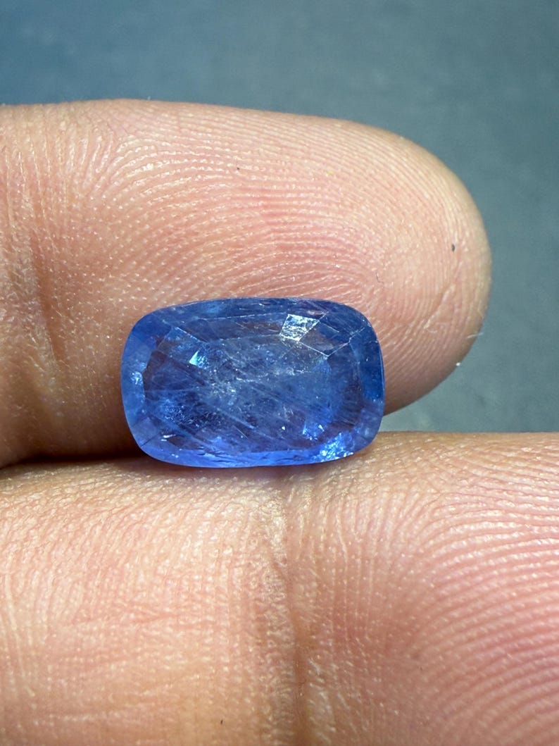 Puede incluir: Una gema azul facetada, de talla coj&iacute;n. La gema se sostiene entre los dedos, mostrando su calidad brillante y transparente. Las facetas de la piedra capturan la luz, creando un efecto brillante. El color general es un azul profundo y vibrante.