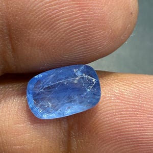 Puede incluir: Una gema azul vibrante con forma rectangular y bordes redondeados. La gema transl&uacute;cida tiene caracter&iacute;sticas internas visibles. La piedra se sostiene entre dos dedos, mostrando su color y textura.