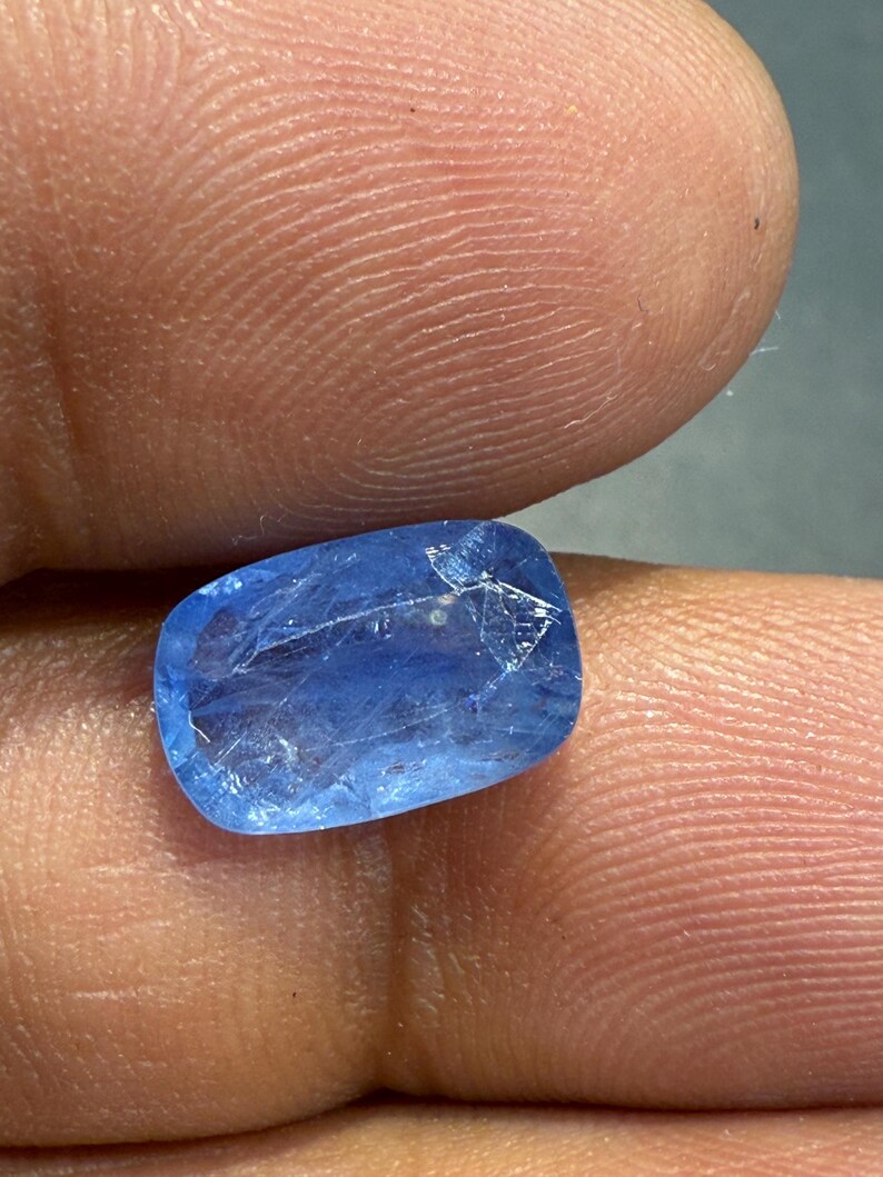 Puede incluir: Una gema azul vibrante con forma rectangular y bordes redondeados. La gema transl&uacute;cida tiene caracter&iacute;sticas internas visibles. La piedra se sostiene entre dos dedos, mostrando su color y textura.
