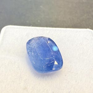 Puede incluir: Una gema azul facetada, de forma ovalada y superficie pulida. La gema se encuentra sobre un fondo blanco, con una superficie gris oscuro en el fondo. La gema tiene una ligera cualidad iridiscente.