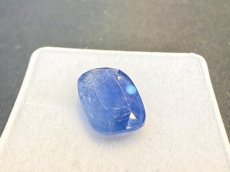 Puede incluir: Una gema azul facetada, de forma ovalada y superficie pulida. La gema se encuentra sobre un fondo blanco, con una superficie gris oscuro en el fondo. La gema tiene una ligera cualidad iridiscente.