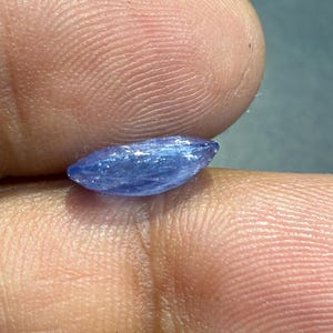 Puede incluir: Una peque&ntilde;a gema azul, alargada, con una textura ligeramente rugosa. La gema se sostiene entre dos dedos, mostrando su forma y color. La piedra parece pulida, reflejando la luz.