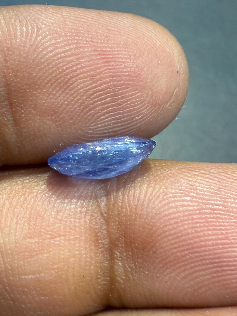 Puede incluir: Una peque&ntilde;a gema azul, alargada, con una textura ligeramente rugosa. La gema se sostiene entre dos dedos, mostrando su forma y color. La piedra parece pulida, reflejando la luz.