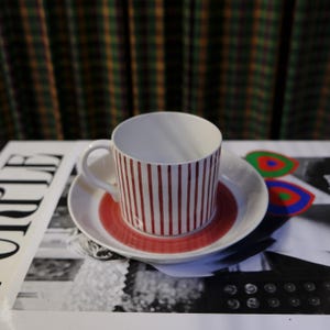 Op de afbeelding: Een witte theekop van keramiek met rode strepen en een bijpassend schoteltje. Het schoteltje heeft een rode ring rond de rand. De theekop en het schoteltje staan op een tijdschrift.