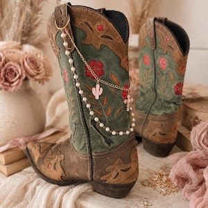 Può includere: Un paio di stivali western marroni e verdi con ricami floreali e sovrapposizioni in pelle marrone. Un accessorio a catena dorata con perle bianche, un piccolo ciondolo a forma di cactus rosa e una croce è attaccato al lato di uno stivale.