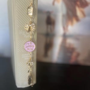 May include: A gold bookmark with several charms attached to it, hanging from a book. The charms include a small rectangle with the word "GOD", angel wings, a pink circle with "Jesus Saves", a cross, and a teardrop charm. The book is a Christian Standard Bible.