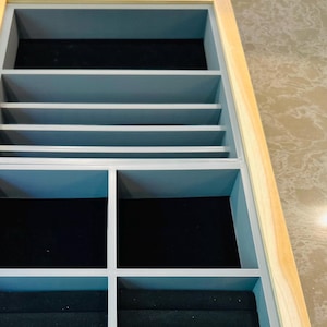 May include: A light blue drawer with a wooden frame and a gold handle. The drawer is open and reveals a gray velvet-lined jewelry organizer with compartments for rings, earrings, and necklaces.