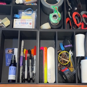 May include: An open drawer with various items organized in compartments. The drawer contains a variety of items, including pens, markers, tape, scissors, a hand sanitizer bottle, nail files, and a lint roller. The drawer is made of wood and has a black liner.
