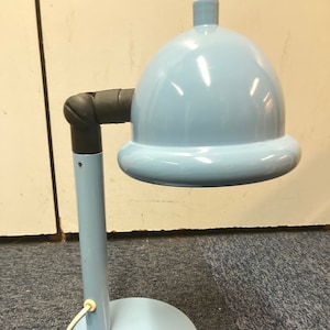 Puede incluir: Una lámpara de escritorio azul claro con una pantalla redonda en forma de cúpula y un brazo negro ajustable. La lámpara tiene un cable blanco y una base redonda.