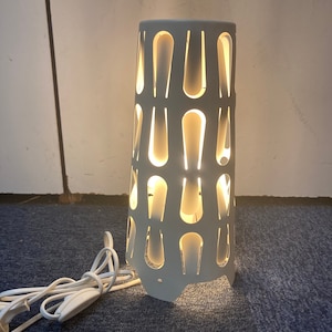 Op de afbeelding: Witte tafellamp met een geometrisch ontwerp. De lamp heeft een wit snoer en een witte schakelaar.