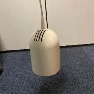 Puede incluir: Lámpara colgante de color crema con una parte superior cónica y una parte inferior cilíndrica redondeada. La parte inferior tiene ranuras verticales para ventilación. Un cable blanco y un cable negro se extienden desde la lámpara.