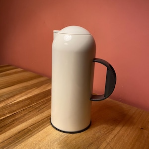 Op de afbeelding: Een cilindrische, gebroken witte thermoskan met een gladde, matte afwerking, een afgerond deksel en een donkergrijs gebogen handvat. De onderkant van de kan is voorzien van een dunne donkergrijze ring die contrasteert met het lichte lichaam.