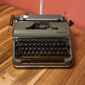 May include: An olive green Olympia typewriter with black keys and chrome accents. The typewriter has a classic design with a carriage return lever and a paper guide. The Olympia logo is visible on the front. The typewriter sits on a wooden surface.