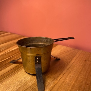 Op de afbeelding: Een kleine, vintage messing pot met een lange handgreep en een standaard. De pot is cilindrisch met een donker interieur en een aangetaste buitenkant. De handgreep en standaard zijn zwart. De pot wordt waarschijnlijk gebruikt om te koken of te verwarmen.