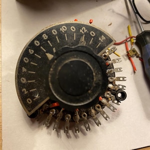 Op de afbeelding: Een zwarte draaischakelaar met een ronde wijzerplaat gemarkeerd met nummers van 0 tot 14. De schakelaar heeft een reeks metalen contacten rond de rand.