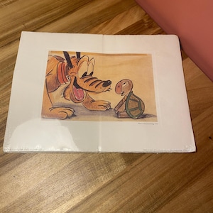 Puede incluir: Una impresión enmarcada con Pluto y una tortuga. Pluto, un perro amarillo con un collar rojo, sonríe a una pequeña tortuga. La impresión está sobre un fondo beige con el título "Pluto's Homecoming 1947".