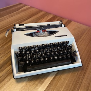 Può includere: Una macchina da scrivere bianca vintage con tasti neri e un carrello cromato. La macchina da scrivere ha un design elegante con una base nera e un accento rosso e bianco sulla parte superiore. I tasti sono disposti in un layout QWERTY standard.