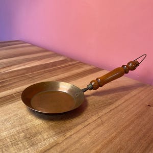 Op de afbeelding: Een kleine, koperkleurige koekenpan met een houten handvat. De pan heeft een rond kookoppervlak en een lange, houten handgreep met een lus aan het uiteinde. De handgreep is aan de pan bevestigd met een metalen fitting. De pan staat op een houten oppervlak.