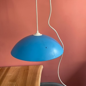 Könnte beinhalten: Eine blaue Pendelleuchte mit weißem Kabel und Deckenhalterung. Die Leuchte hat einen abgerundeten, schalenförmigen Schirm. Der Hintergrund ist einfarbig korallenfarben.