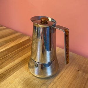 Op de afbeelding: Een roestvrijstalen espressopot voor op het fornuis met een taps toelopend cilindrisch lichaam. Het deksel heeft een kleine, ronde, amberkleurige gespikkelde knop en het handvat aan de zijkant is voorzien van een bijpassende amberkleurige inleg.