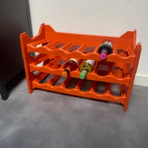 Op de afbeelding: Een feloranje modulair wijnrek van kunststof met drie niveaus van golvende flessenhouders. Het rek bevat drie glazen flessen met verschillend gekleurde doppen, waaronder één donkere fles op het bovenste niveau en twee op het middelste.