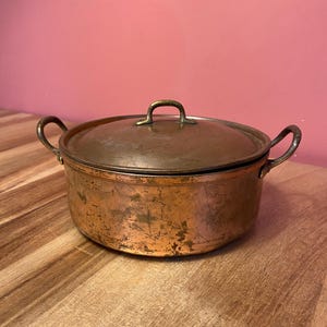 Op de afbeelding: Een koperen pot met een deksel, met twee handvatten aan de zijkant en een klein handvat op het deksel. De pot heeft een verweerde afwerking, met tekenen van ouderdom en gebruik. De pot staat op een houten oppervlak.