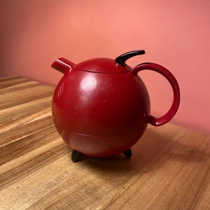 Puede incluir: Una tetera esférica de color rojo con acabado liso, que cuenta con un asa negra, un pomo negro en la tapa y tres pequeñas patas negras. La tetera tiene un pico corto e inclinado en el lateral y un asa curva unida al cuerpo principal.