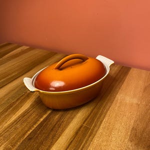 May include: An orange oval-shaped ceramic casserole dish with a matching lid and a curved handle. The dish has white handles on each side. The dish is on a wooden surface.