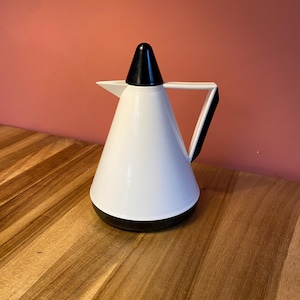 Op de afbeelding: Een witte en zwarte thermoskan met een puntige deksel en een handvat. De kan staat op een houten oppervlak.