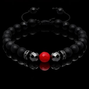 Puede incluir: Una pulsera de cuentas negras con una cuenta roja central y dos cuentas grises facetadas. La pulsera tiene un cierre de cordón negro ajustable. Las cuentas están sobre un fondo negro.