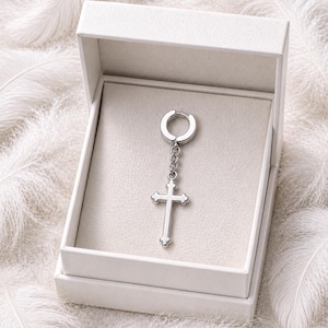 May include: A silver-colored cross earring with a hinged hoop, displayed in a white box. The cross hangs from a chain and has a detailed design. The earring is set against a backdrop of white feathers.