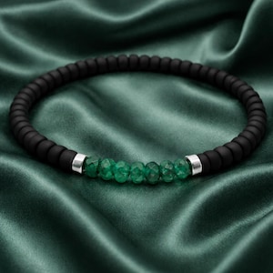 Puede incluir: Pulsera de cuentas negras con una sección de cuentas verdes esmeralda facetadas. La pulsera tiene detalles metálicos plateados. El fondo es una tela de seda verde oscuro.