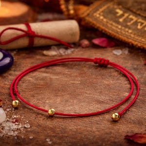Puede incluir: Una pulsera de hilo rojo con dos cuentas doradas, exhibida sobre una superficie de madera. La imagen incluye una vela encendida, un pergamino atado con una cuerda roja, un amuleto azul y blanco en forma de ojo y pétalos de rosa esparcidos.