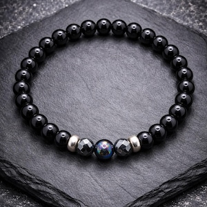 Op de afbeelding: Een kralenarmband met gladde, ronde zwarte kralen in een cirkelvormig patroon. Het midden bevat twee zilverkleurige metalen tussenstukjes, twee gefacetteerde donkergrijze kralen en één centrale iriserende kraal met een donkere, reflecterende afwerking.
