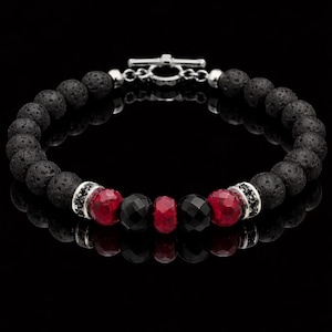 Puede incluir: Una pulsera de cuentas de piedra de lava negra con detalles de piedras preciosas rojas y negras. La pulsera presenta un cierre de palanca plateado y se muestra sobre una superficie negra reflectante. Las cuentas tienen aproximadamente 0,8 cm de diámetro.
