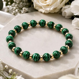 Puede incluir: Una pulsera de cuentas que alterna esferas de malaquita verde con bandas oscuras características y pequeñas cuentas espaciadoras de metal dorado pulido, dispuestas en un patrón circular para formar un accesorio flexible.