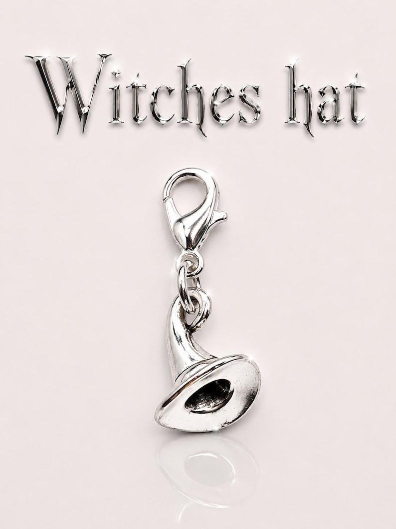May include: A silver-colored charm in the shape of a witch's hat, with a lobster clasp. The word "Witches hat" is above the charm. The charm is on a light pink background.