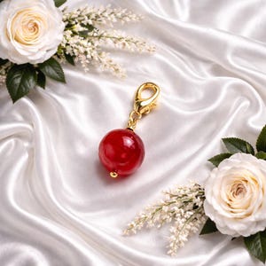 May include: A red, spherical charm with a gold-coloured clasp and detailing. The charm is set against a backdrop of white satin fabric, with white roses and delicate white flowers in the corners.