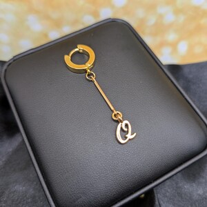 Può includere: Un orecchino color oro con una barra pendente e un ciondolo con la lettera "Q" in corsivo. L'orecchino ha una chiusura a cerchio ed è esposto su una superficie nera.