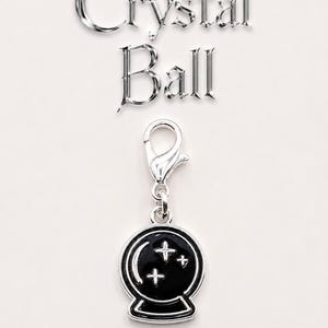 May include: A silver-colored charm with a black crystal ball design, featuring three white stars inside. The charm is attached to a silver clasp. The words "Crystal Ball" are displayed above the charm in a metallic font.
