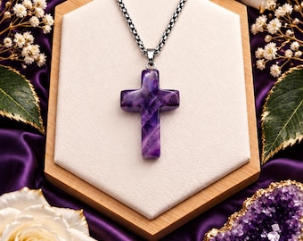 May include: A purple amethyst cross pendant suspended from a silver-toned metal box chain. The cross features natural deep violet and white banding, showcasing a smooth, polished finish. The necklace is displayed on a light-colored hexagonal base.
