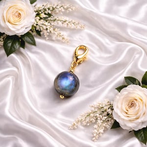 May include: A gold-colored charm with a round, iridescent blue and gray stone. The charm is attached to a gold clasp. The background is white satin fabric with white roses and small white flowers.