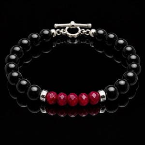 Könnte beinhalten: Armband mit schwarzen Onyxperlen und einer Reihe facettierter roter Perlen. Es hat einen silbernen Knebelverschluss. Die Perlen haben einen Durchmesser von etwa 8 mm. Das Armband ist ein Schmuckstück.